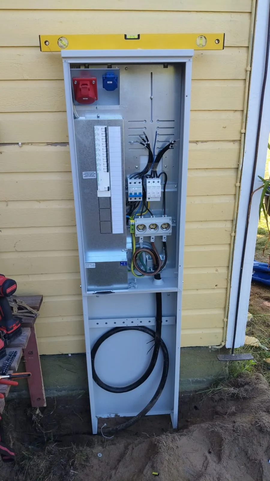 Asentamaton sähkökaappi keltaisen talon seinää vasten, yläosassa vesivaaka tasossa.