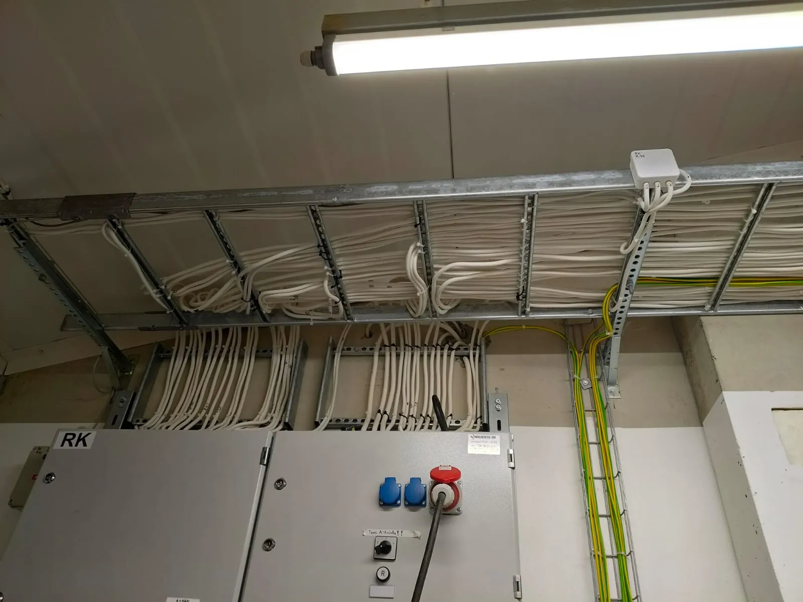 Sähkökeskus ja järjestelmällisesti asennetut valkoiset ja keltaiset sähköjohdot metalliverkossa katossa.